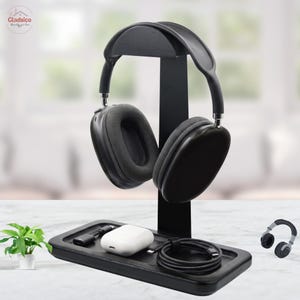 May include: A black headphone stand with a rectangular base tray. The stand features a curved top support for over-ear headphones. The base tray holds a white wireless earbud charging case, a black charging cable, and a small black adapter.