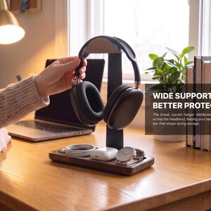 May include: A dark wood headphone stand with a black vertical support and a curved top hanger. The base features a tray holding a white charging cable, white wireless earbuds in a case, and a silver wristwatch. Text reads: WIDE SUPPORT FOR BETTER PROTECTION.