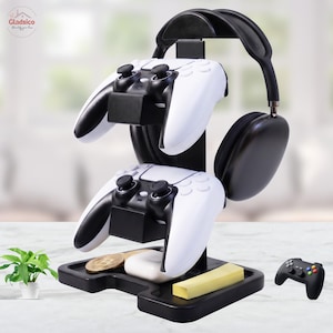 May include: A black gaming accessory stand featuring two tiers for holding white and black game controllers, a top arch for over-ear headphones, and a base tray containing a small white case and a yellow rectangular block.