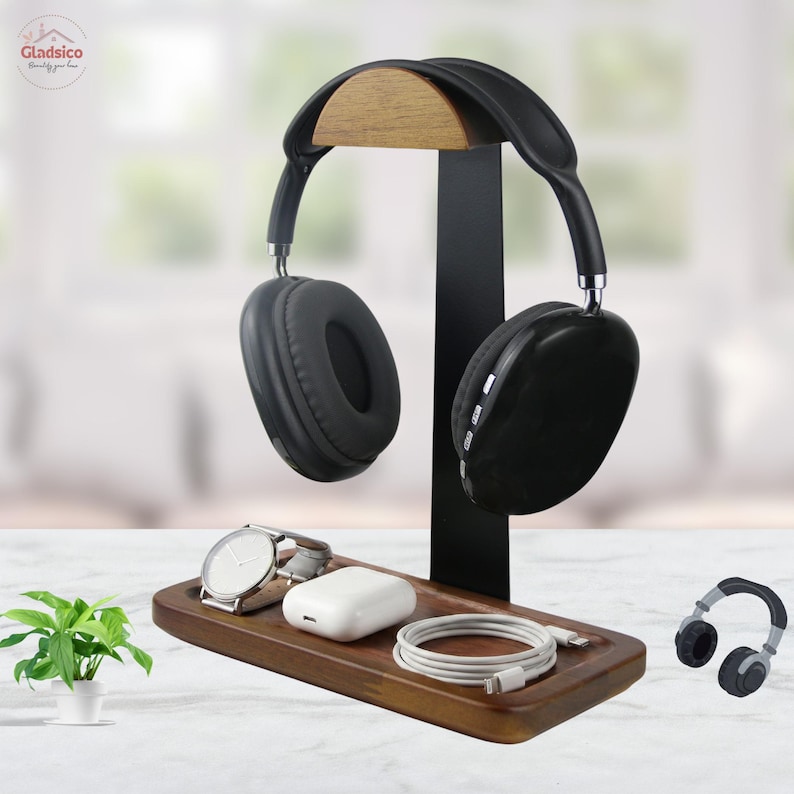 May include: A wooden headphone stand with a black metal vertical support and a curved wooden top rest. The base is a rectangular wooden tray holding a silver wristwatch, a white charging case, and a coiled white charging cable.