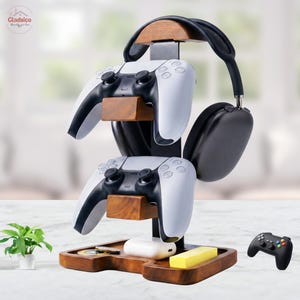 May include: A multi-tiered wooden and black metal stand designed to hold two white and black game controllers and a pair of black over-ear headphones. The base features a wooden tray with a small white charging case and a yellow rectangular block.