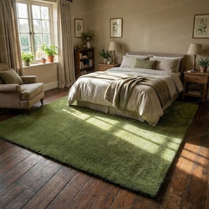May include: A bedroom scene featuring a large, green, rectangular rug. The rug is placed on a dark wooden floor, with a bed, armchair, and window visible in the background. The room is decorated in neutral tones, creating a cozy atmosphere.