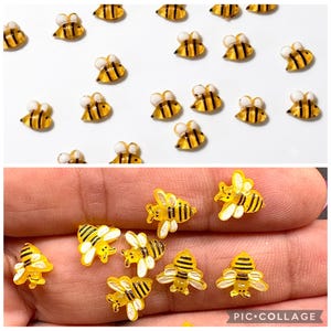 May include: A collection of small, flat-backed bee-shaped embellishments. Each bee features a translucent yellow body with black horizontal stripes and opaque white wings. The bees are arranged in a scattered pattern and also shown in a group.