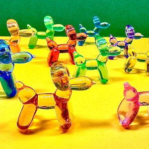 May include: A collection of colorful glass balloon dog figurines in various colors, including blue, green, red, yellow, and pink. The dogs are arranged on a yellow surface with a green background, creating a vibrant and playful display.