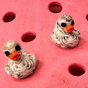 Op de afbeelding: Twee kleine, decoratieve eendenbedels met zwarte ogen en oranje snavels. De witte eenden hebben bruine details en liggen op een roze ondergrond met ronde gaten. Deze bedels zijn waarschijnlijk schoenaccessoires.