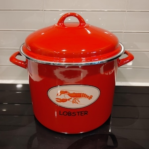 May include: A bright red enamel stockpot with a matching lid and two side handles. The front features an oval cream-coloured decal with an orange lobster illustration and the word LOBSTER printed in black lettering below the graphic.