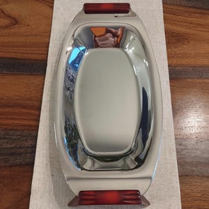 May include: A polished, oval serving tray with a reflective stainless steel surface. The tray has dark red, rectangular handles at each end. The tray is designed for serving food and drinks.