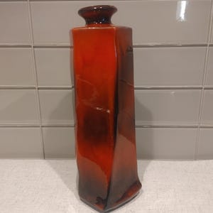 Peut inclure: Un grand vase en céramique rectangulaire avec une finition brillante. Le vase présente un dégradé de couleurs vives, passant d'un brun foncé profond à la base à un orange vif et ardent vers le haut, se terminant par un col étroit et arrondi.
