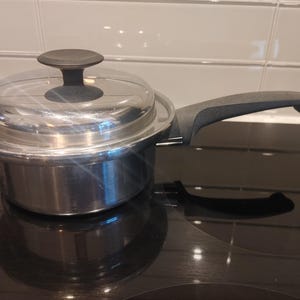 May include: A stainless steel saucepan with a clear glass lid and a black handle. The lid features a central black knob. The pan has a polished metallic finish and a long, ergonomic black handle extending from the side, designed for stovetop use.