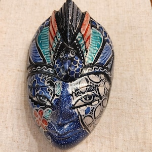 May include: A hand-carved wooden mask featuring a central bird figure at the top with red, teal, and black accents. The face is painted in shades of blue, white, and black with intricate floral and geometric patterns, including a prominent red flower.