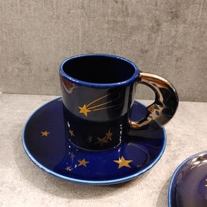 May include: A dark blue ceramic coffee cup and saucer set. The cup has a gold handle and is decorated with gold stars and a shooting star design. The saucer also features gold star accents. The set is designed for coffee or tea.