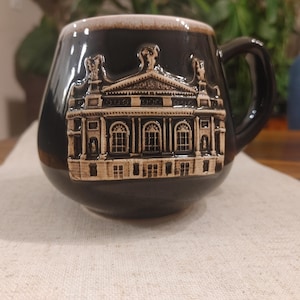 May include: A dark brown ceramic mug featuring a raised, light-coloured relief sculpture of a classical building with columns and arched windows on its front. The mug has a rounded body, a curved handle, and a light-coloured rim at the top edge.