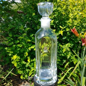 Peut inclure: Une carafe en verre transparent avec une base carrée et un bouchon en verre assorti. La carafe présente un corps haut et rectangulaire aux coins arrondis et une base épaisse et lourde. Une petite étiquette carrée est visible sur le verre.