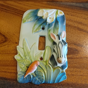 Op de afbeelding: Een rechthoekige keramische lichtschakelaarplaat met een verhoogd, driedimensionaal ontwerp van een zebrahoofd aan de rechterkant, een kleine oranje vogel linksonder en witte bloemen met groene bladeren op de achtergrond in een glanzende afwerking.