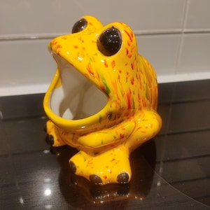 Op de afbeelding: Een keramieken kikkerbeeldje met een wijd open mond, voorzien van een felgele glazuurlaag met gespikkelde rode en groene verfaccenten. De kikker heeft grote, glanzende zwarte ogen en donkerbruin geschilderde tenen.