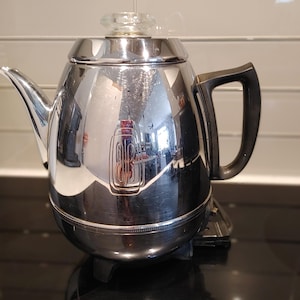 May include: A polished chrome electric percolator coffee pot featuring a black handle, a clear glass knob on the lid, and a prominent embossed logo on the front. The metallic surface reflects the surrounding environment, and it sits on a black base.