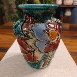 May include: A ceramic vase featuring a glossy glaze in shades of teal, deep red, and cream. The surface is decorated with raised, embossed floral motifs, including a prominent light blue flower with a yellow centre and textured leaf patterns.