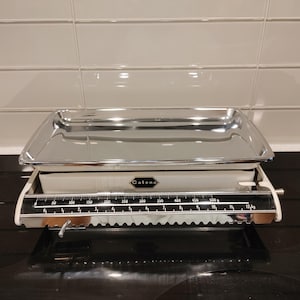 May include: A white Catena kitchen scale featuring a large, rectangular, polished chrome tray on top. The front display shows a black scale with white markings, measuring up to 12 kg and 500 grams, with a clear, horizontal sliding indicator.