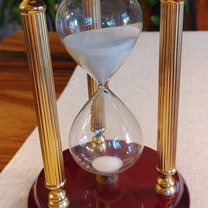 May include: An hourglass featuring a clear glass bulb filled with white sand, supported by three vertical gold-toned fluted metal pillars. The top and bottom circular bases are crafted from a polished dark reddish-brown wood with a glossy finish.