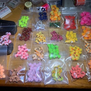 May include: Assortment of colorful fly tying materials in individual plastic bags. The materials are arranged in rows and include various colors of fur, feathers, and beads.
