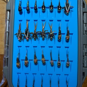 May include: A blue fly fishing fly box containing rows of different coloured flies. The flies are tied with various materials, including feathers, beads, and thread.