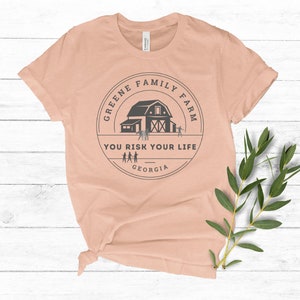 May include: Peach colored t-shirt with a black and white graphic of a barn with the text "Greene Family Farm You Risk Your Life Georgia".