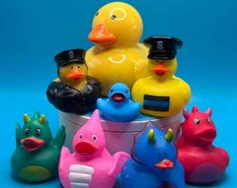 May include: A collection of eight rubber duck toys, including a yellow duck with a black beak, a blue duck, a pink duck, a green duck, a red duck, a blue duck with green horns, a black duck with a police hat, and a yellow duck with a police hat and a blue stripe.