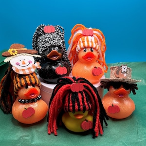 May include: Five rubber duckies decorated for fall with various yarn hair styles, hats, and pumpkin accents. The ducks are sitting on a green surface.