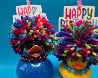 May include: Two rubber duckies with colorful yarn hair and birthday hats that say "Happy Birthday". One duck is blue with red beads and sunglasses, the other is yellow with blue beads.