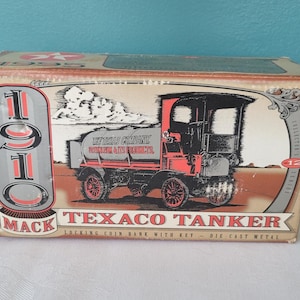 Op de afbeelding: Een vintage-stijl spaarpot van gegoten metaal in de vorm van een Mack Texaco tankwagen uit 1910. De doos heeft een rood, wit en zwart voertuig met de tekst "The Texas Company Gasoline & Its Products" aan de zijkant van de tank. De doos vermeldt ook "Locking Coin Bank with Key - Die Cast Metal" en "Collector's Edition #12".