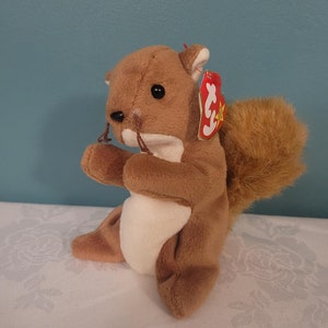 May include: A brown plush squirrel toy with a fluffy tail and a red tag. The squirrel has black eyes and is holding a small object in its paws. The toy is sitting on a white surface.
