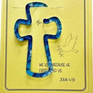 Peut inclure: Marque-page en forme de croix en vitrail bleu avec un motif floral et une illustration de colombe sur une carte jaune. La carte porte le texte "WE LOVED BECAUSE HE FIRST LOVED US. JOHN 4:19."