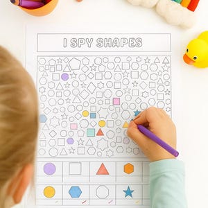 May include: A child's hand using a purple marker to color a shape on an "I Spy Shapes" activity sheet. The sheet features various shapes and a key at the bottom. Crayons, a star, a rainbow, and a rubber duck are also visible.