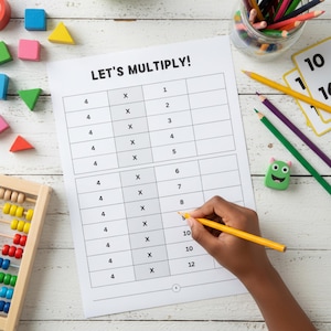 May include: A worksheet titled "LET'S MULTIPLY!" with multiplication problems is shown. A hand holding a yellow pencil is working on the worksheet. Colored pencils, erasers, and number cards are scattered around the worksheet. An abacus and wooden blocks are also visible.