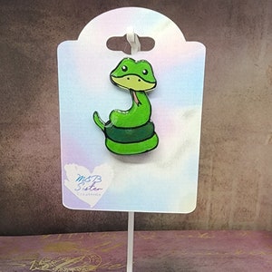 May include: Green snake enamel pin with a white background. The pin is on a white display stand with a heart-shaped sticker that says "M&B Sister Creations".