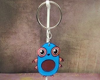May include: Blue and brown cartoon creature keychain with large eyes and a smiling mouth. The keychain is attached to a silver ring.