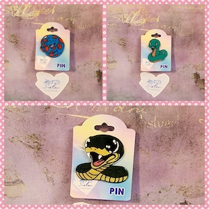 Op de afbeelding: Drie emaillepins met verschillende slangontwerpen. De pins bevinden zich op een witte achtergrond met een roze rand. De tekst "PIN" is op elke kaart gedrukt. De tekst "M&B Sister Creations" is op elke kaart gedrukt.