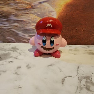 May include: A pink and red figurine of a character wearing a red cap with a white "M" on it. The character has a smiling face with blue eyes and a brown moustache.