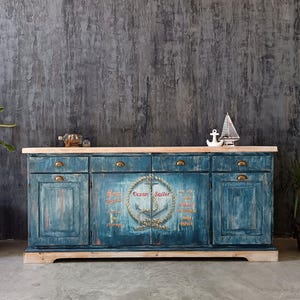 Könnte beinhalten: Ein langes, rechteckiges Sideboard aus Holz mit einem abgenutzten blauen Finish und einer naturfarbenen Holzplatte. Es verfügt über vier Schubladen mit Messinggriffen und zwei Schränke. Die mittleren Türen zeigen eine Ankergrafik mit dem Text Ocean Sailor.