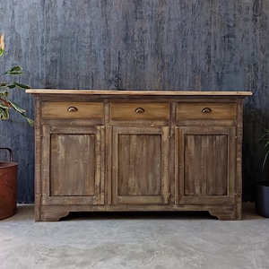 Peut inclure: Un buffet rectangulaire en bois avec un plateau en bois naturel et une finition brune vieillie. Le meuble comprend trois tiroirs supérieurs avec de petites poignées en métal et trois portes de placard inférieures, chacune avec un panneau central.