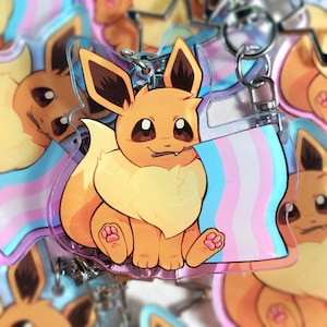 May include: A clear acrylic keychain featuring a cartoon Eevee character with a transgender pride flag. The Eevee is orange and yellow with brown accents, holding the flag with blue, pink, and white stripes. The keychain has a silver metal ring.