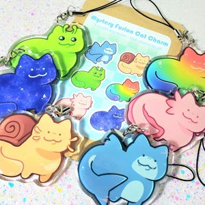 May include: A collection of colorful acrylic keychains featuring cartoon cat designs. The keychains include various colors like green, blue, pink, and yellow, with some featuring galaxy patterns and rainbow gradients. A card with the text "Mystery Fusion Cat Charm" is also visible.