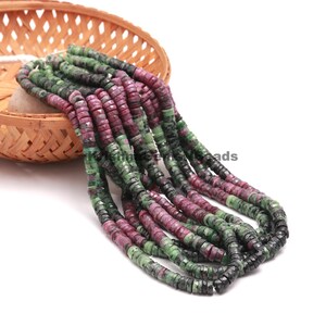 May include: Strands of gemstone beads in shades of green and burgundy are displayed. The beads are disc-shaped and appear to be made of natural stone. They are arranged in a cascading pattern, with some beads resting in a woven basket.
