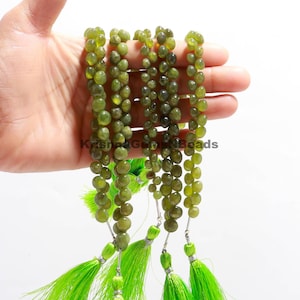以下が含まれることがあります： オリーブグリーンの宝石ビーズの複数のストランドと明るいグリーンのタッセル。ビーズは丸く、一緒にひもでつながっています。タッセルはシルバーカラーの金属で取り付けられています。「Krishna Gems Beads」というテキストが画像に表示されています。