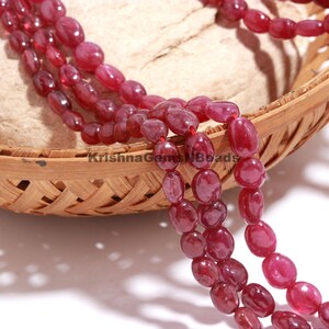 Peut inclure: Un collier de perles de rubis rouge foncé de forme ovale enfilées ensemble. Les perles ont une finition lisse et polie et sont drapées sur un panier en bambou tressé. Le texte KrishnaGemsNBeads est imprimé en noir sur le bord du panier.