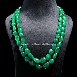 Può includere: Una collana a tre fili con grandi perle verdi ovali lucide. La collana è esposta su un manichino nero. Il testo "KrishnaGemsNBeads" è visibile.