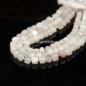 May include: A strand of white moonstone beads, each bead is faceted and has a square shape. The beads are strung on a white thread.