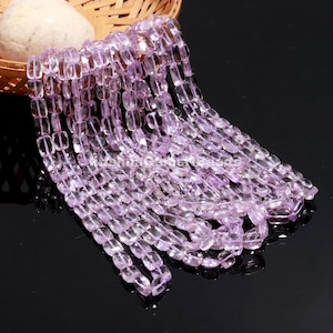 May include: Strands of light purple, rectangular beads cascade from a woven basket. The beads are translucent, with a subtle sheen, and appear to be made of a polished stone or crystal. The beads are strung together, creating a flowing, elegant display.