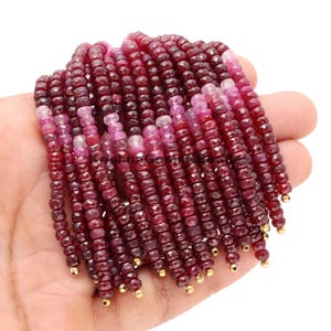 May include: Strands of faceted ruby beads in varying shades of red and pink. The beads are small and uniform in size, with gold-colored end caps. The beads are strung together, creating a cascading effect.