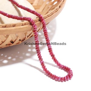 May include: A strand of small, round, deep pink ruby beads arranged in a curved line. The beads have a smooth, polished texture and a consistent size. The text KrishnaGemsNBeads is visible in the center of the image, positioned near the beads.